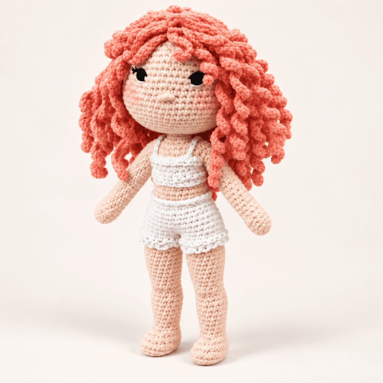 Ghita, a hand-crocheted doll with curly coral hair, a white crochet camisole and ruffled shorts, standing against a soft cream backdrop.